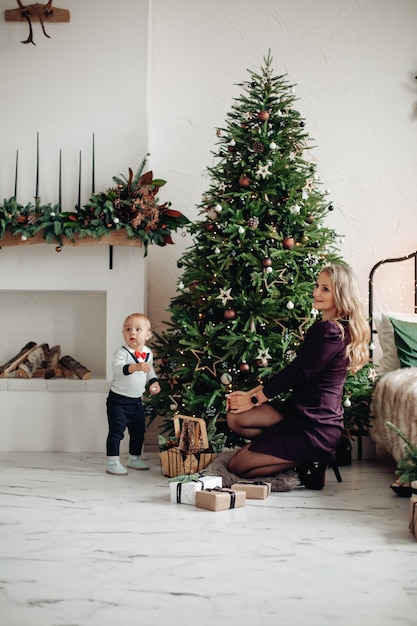 Linda loira mãe e lindo filho ao lado da árvore de Natal decorada em casa. Celebração familiar.