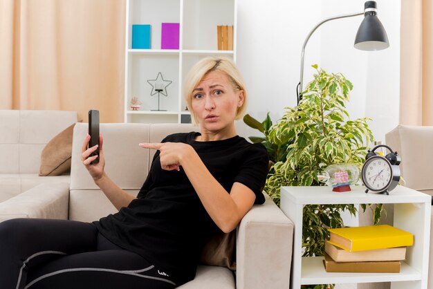 Linda loira mulher russa confusa deitada na poltrona segurando e apontando para o telefone dentro da sala de estar