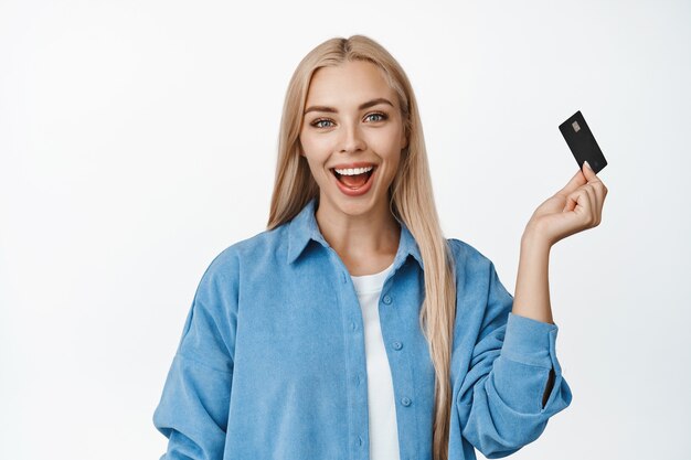 Linda loira parecendo espantada, mostrando seu cartão de crédito, conceito de compras online e pagamento sem contato em branco.