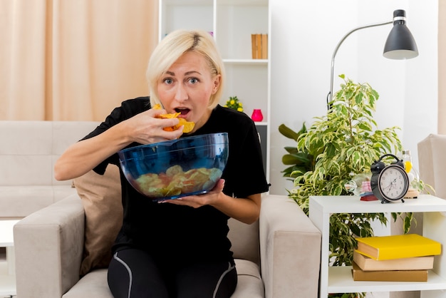 Linda loira russa animada sentada na poltrona segurando e comendo uma tigela de batatas fritas dentro da sala de estar