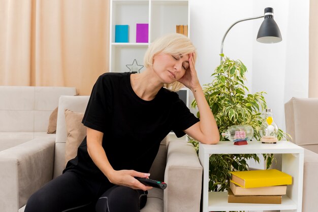 Linda loira russa com sono sentada na poltrona colocando a mão na cabeça segurando o controle remoto da TV na sala