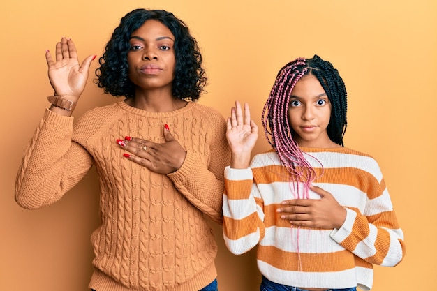 Foto grátis linda mãe afro-americana e filha vestindo suéter de inverno de lã xingando com a mão no peito e palma da mão aberta fazendo um juramento de promessa de lealdade