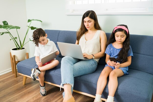 Linda mãe digitando no laptop e trabalhando enquanto está sentada no sofá com seu filho e filha. Crianças brincando com um tablet e smartphone