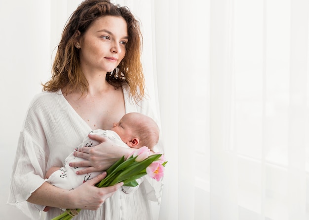 Foto grátis linda mãe olhando para longe enquanto carregava seu bebê em pé perto de cortina branca