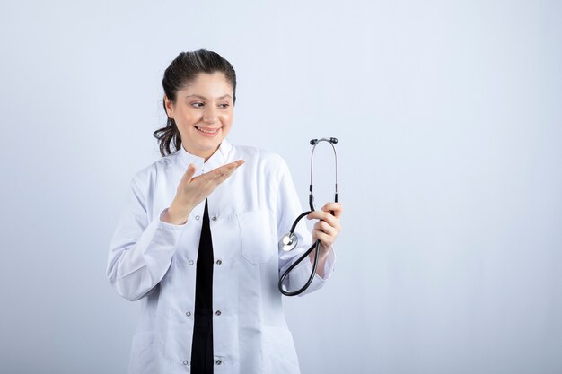 Linda médica de jaleco branco segurando o estetoscópio e sorrindo.