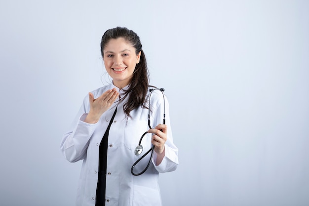 Linda médica de jaleco branco segurando o estetoscópio e sorrindo.