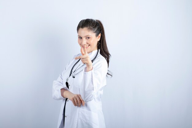Linda médica feminina em jaleco branco posando com estetoscópio sobre uma parede branca.