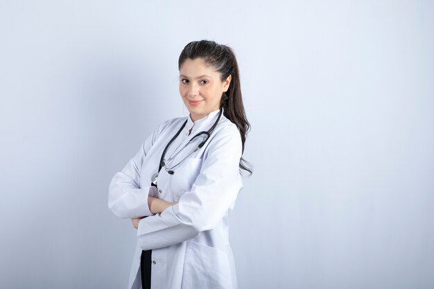 Linda médica nos braços eretos de jaleco branco cruzados sobre uma parede branca.