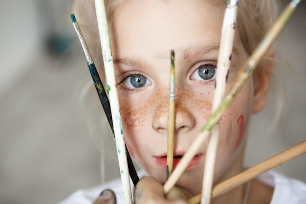 Linda menina loira com grandes olhos azuis, posando com pincéis dentro de casa. Menina brincando minúscula, apreciando a pintura.