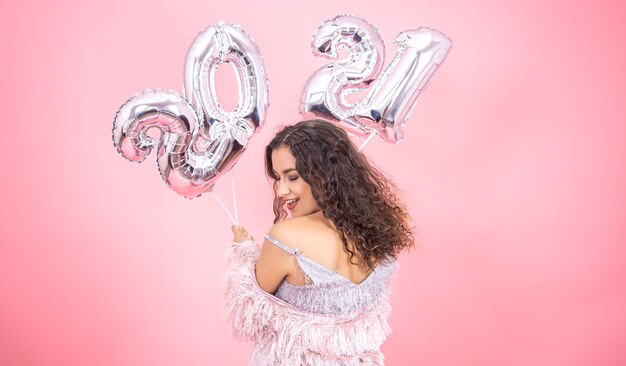 Linda menina morena com ombros nus em roupas festivas, posando em um fundo rosa com balões para o ano novo em suas mãos