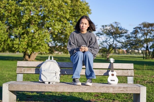 Linda menina morena no banco no parque sentado com ukulele e mochila segurando smartphone usando aplicativo móvel