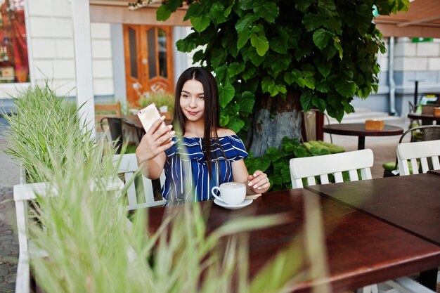 Linda menina morena sentada na mesa no café com uma xícara de café e fazendo selfie