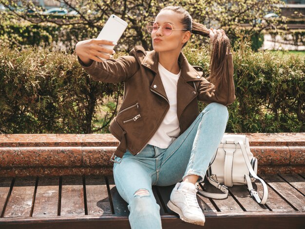 linda menina morena sorridente no verão hipster jaqueta e calça jeans modelo tomando selfie em smartphone