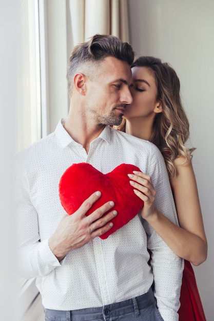 Linda moça de vestido vermelho beijar seu homem e segurando o coração de brinquedo de pelúcia