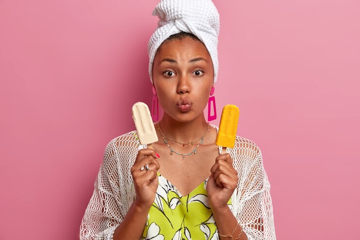 Foto grátis linda modelo de pele escura mantém os lábios dobrados, posa com sorvetes de baunilha e manga, faz refeições não saudáveis durante o verão, usa toalha de banho e vestido casual, modelos contra a parede rosa