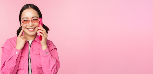 Linda modelo feminina coreana em óculos de sol falando no celular com cara de feliz usando o serviço de celular para ligar para o amigo no smartphone em pé sobre fundo rosa