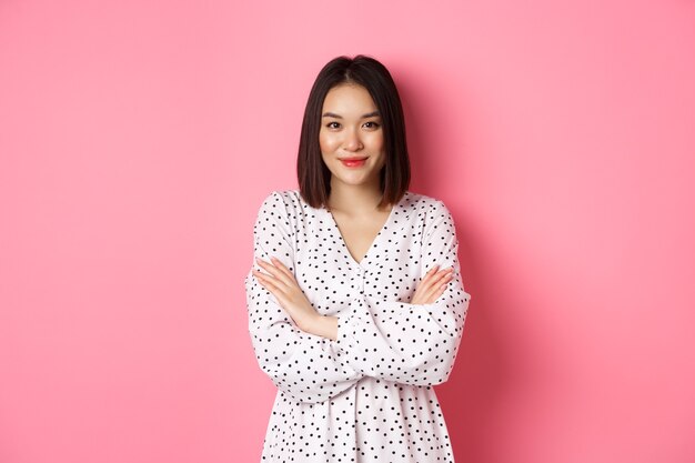 Linda modelo feminino asiático em pé no vestido de braços cruzados no peito e sorrindo para a câmera em pé ov ...