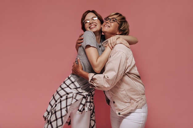 Linda morena com óculos em t-shirt cinza, calça branca e camisa xadrez, abraçando uma mulher loira sorridente em uma jaqueta bege em pano de fundo rosa.