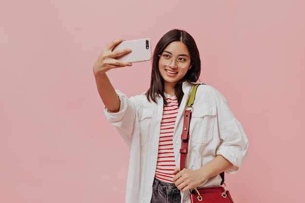 Linda morena de camiseta listrada e óculos segura bolsa vermelha Linda mulher de camisa branca tira selfie em fundo rosa