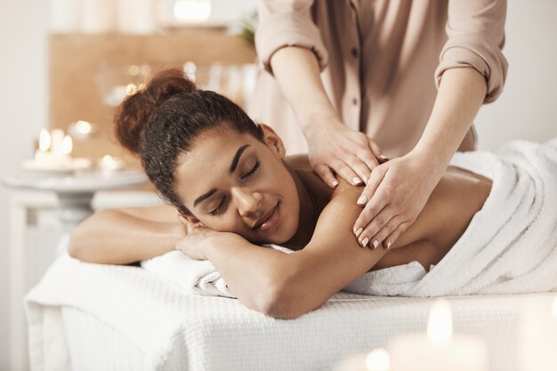 Linda mulher africana sorrindo desfrutando de massagem com os olhos fechados no salão spa.