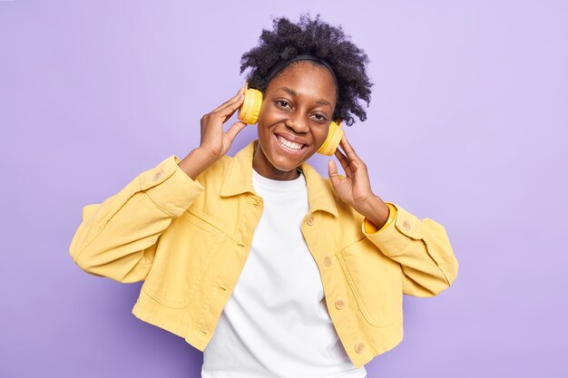 Linda mulher afro-americana de pele escura positiva com sorriso cheio de dentes ouve música por meio de fones de ouvido sem fio e desfruta do tempo livre inclina a cabeça