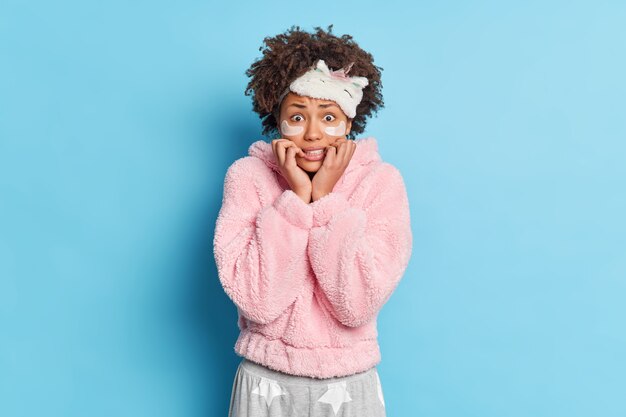 Linda mulher afro-americana preocupada com cabelo encaracolado olha nervosamente para a câmera, vestida com pijamas e usa máscara de dormir na testa isolada sobre a parede azul