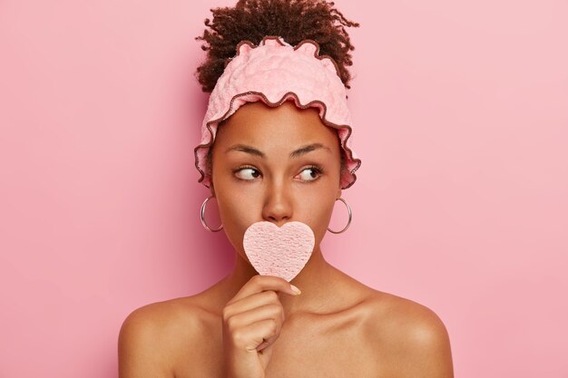 Linda mulher afro mantém a pele em boas condições, tenta relaxar e se desestressar, remove a sujeira do dia a dia com esponja cosmética, desvia o olhar, usa bandana de chuveiro rosa e brincos redondos