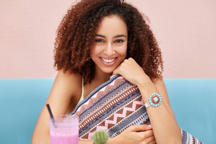 Foto grátis linda mulher alegre com penteado afro, segura uma almofada macia, se diverte sozinha em um restaurante aconchegante, gosta de uma bebida de verão, sorri com alegria, senta-se contra a parede rosa.