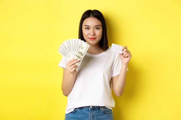 Linda mulher asiática com cabelo escuro curto, vestindo camiseta branca, mostrando dinheiro em dólares e cartão de crédito de plástico, em pé sobre fundo amarelo.