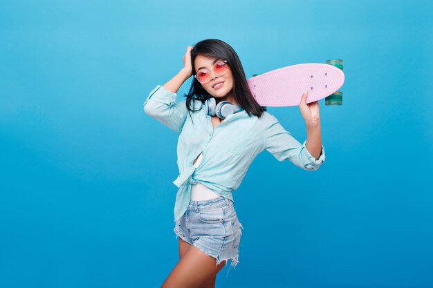 Linda mulher asiática com cabelo liso usa shorts jeans, posando com o skate na sala com interior luminoso. retrato de garota hispânica confiante em óculos de sol bonitos desfrutando.