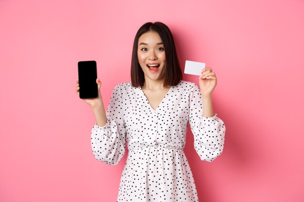 Linda mulher asiática, compras online, mostrando o cartão de crédito do banco e a tela do celular, sorrindo e olhando para c ...