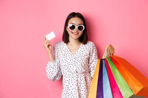 Linda mulher asiática em óculos de sol, vai às compras, segurando sacolas e mostrando o cartão de crédito em pé ...