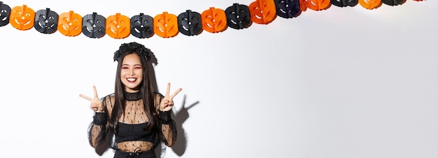 Linda mulher asiática feliz curtindo a festa de halloween mostrando gesto de paz e sorrindo usando custo de bruxa