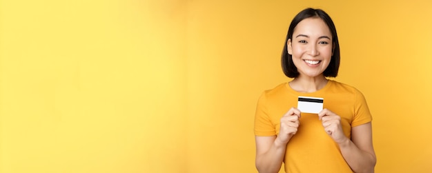 Linda mulher asiática mostrando cartão de crédito e sorrindo recomendando serviço bancário em pé sobre amarelo...