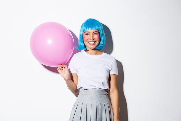 Linda mulher asiática na peruca azul e fantasia de colegial para o halloween, segurando o balão rosa e sorrindo, em pé.