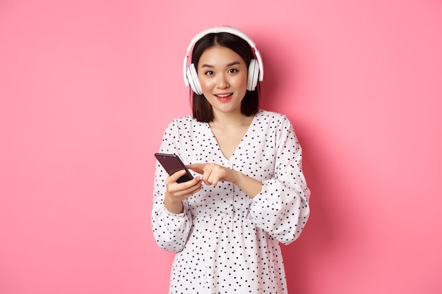 Linda mulher asiática ouvindo música em fones de ouvido, usando o celular, sorrindo feliz para a câmera, em pé sobre um fundo rosa