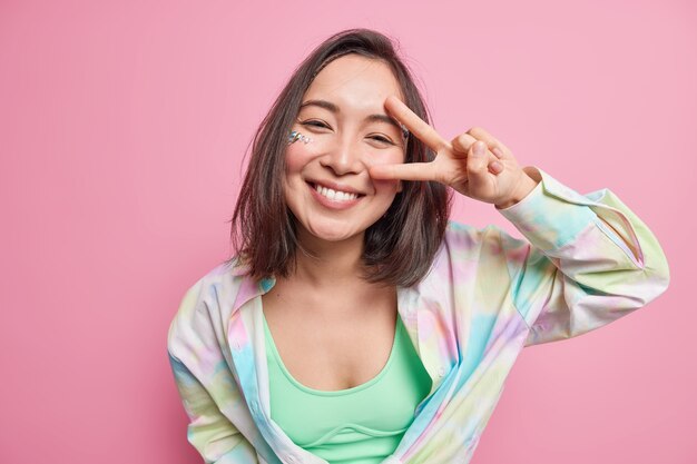 Linda mulher asiática positiva com cabelo escuro mostra sinal de v gesto de paz parece alegremente vestida com camisa casual goza de bom dia isolado sobre a parede rosa expressa emoções despreocupadas.