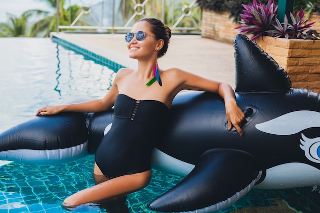 Linda mulher asiática se divertindo na piscina em uma vila tropical nas férias de verão na Tailândia, brincando com uma grande orca vestindo maiô preto e óculos escuros, corpo sexy, acessórios de moda