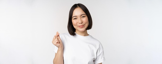 Linda mulher asiática sorrindo mostrando gesto de coração de dedo vestindo camiseta em pé contra ba branco