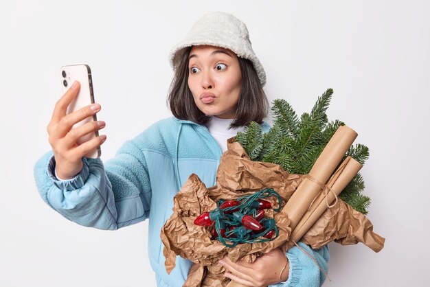 Linda mulher asiática tira selfie via smartphone mantém lábios arredondados envia vibrações positivas com interlocutor se comunicando on-line se prepara para poses de ano novo com itens de decoração poses internas