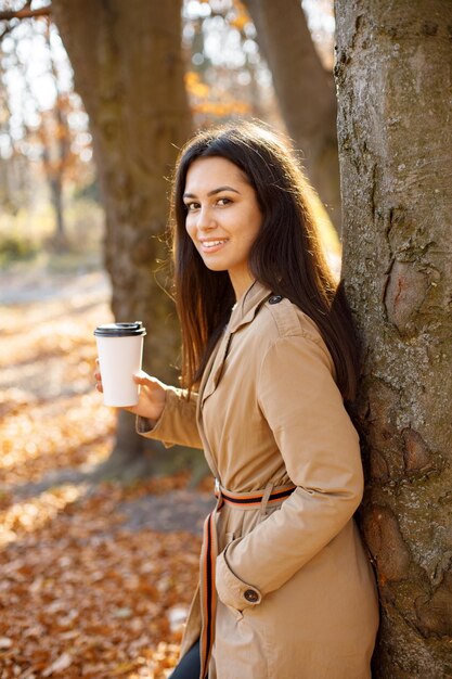 Linda mulher bebe café e posando para a câmera no parque outono. Jovem de pé perto de árvore com um café. Mulher morena vestindo casaco bege.
