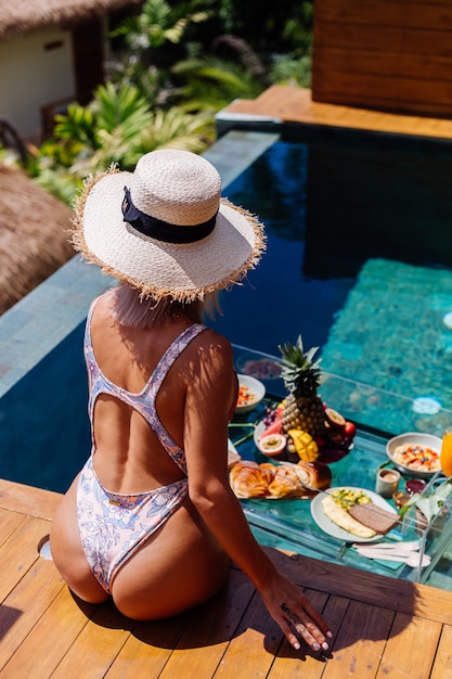 Linda mulher bronzeada caucasiana de biquíni e chapéu de palha com café da manhã flutuante na villa de estilo bali de luxo incrível em dia de sol na piscina, fundo tropical.