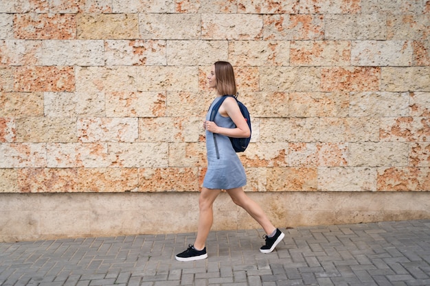 Linda mulher caminhando com uma mochila em vestido azul caminhando pela cidade em parede decorada com pedra com textura marinha