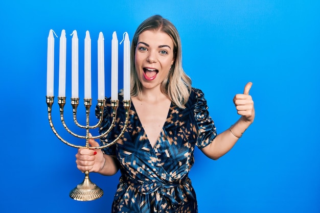 Linda mulher caucasiana segurando menorah hanukkah vela judaica apontando o polegar para o lado sorrindo feliz com a boca aberta