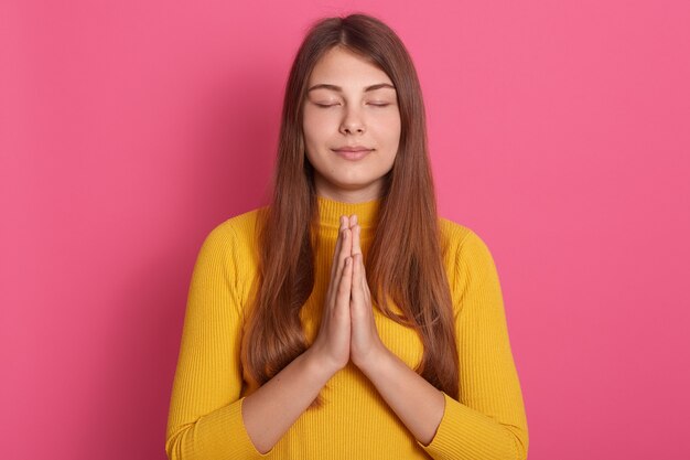 Linda mulher com cabelo comprido rezando com os olhos fechados, mantendo as palmas das mãos unidas, posando isolado no espaço rosa, vestindo um suéter amarelo