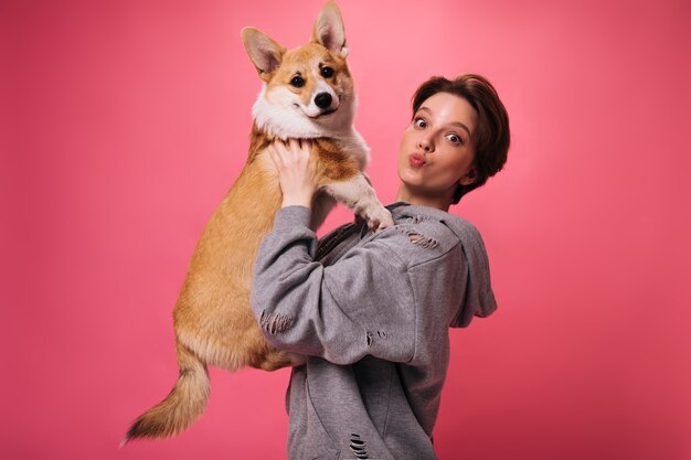 Linda mulher com capuz detém cachorro no fundo rosa. Mulher charmosa de cabelos escuros e roupa cinza brincando com corgi isolado