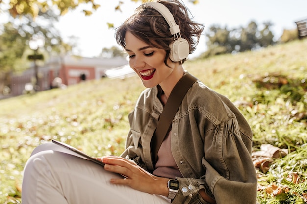 Foto grátis linda mulher com jaqueta e fones de ouvido, sentado na grama ao ar livre. mulher feliz com penteado encaracolado, segurando o smartphone do lado de fora.