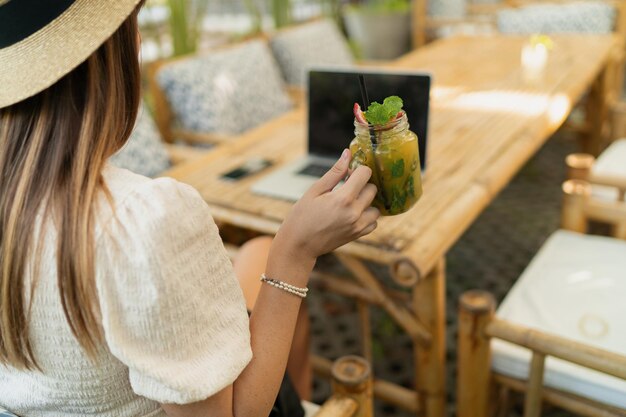 Linda mulher de chapéu de palha usando lap top enquanto viaja na ásia Trabalhando remotamente em elegante café tropical Segurando bebida exótica