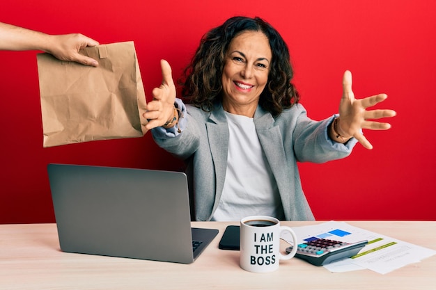 Foto grátis linda mulher de meia-idade trabalhando no escritório pedindo comida de entrega olhando para a câmera sorrindo de braços abertos para o abraço. expressão alegre abraçando a felicidade.