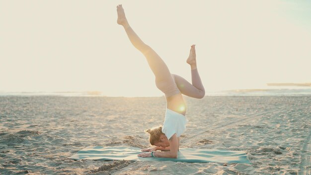 Linda mulher elegante fazendo pose de ioga parada de mãos com divisões na praia vazia Jovem professora de ioga deslumbrante praticando ioga no tapete